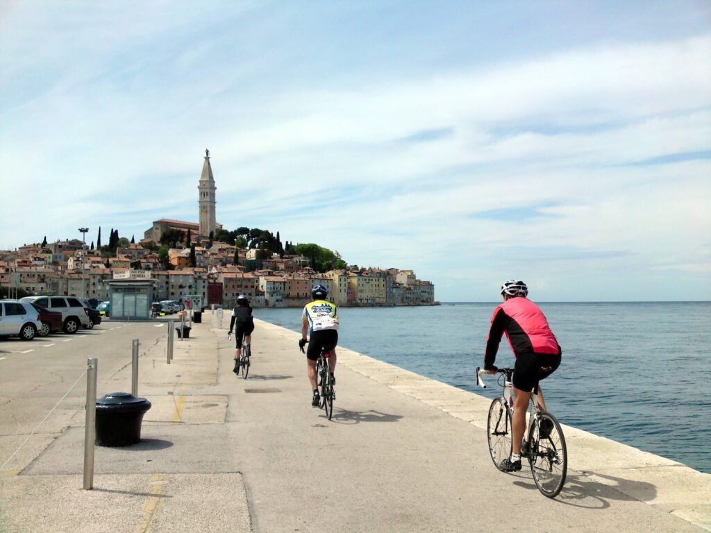 Rennradreise Kroatien Istrien, drei Rennradler fahren entlang einer Kaimauer an der Adria auf die schöne Hafenstadt Rovinj zu