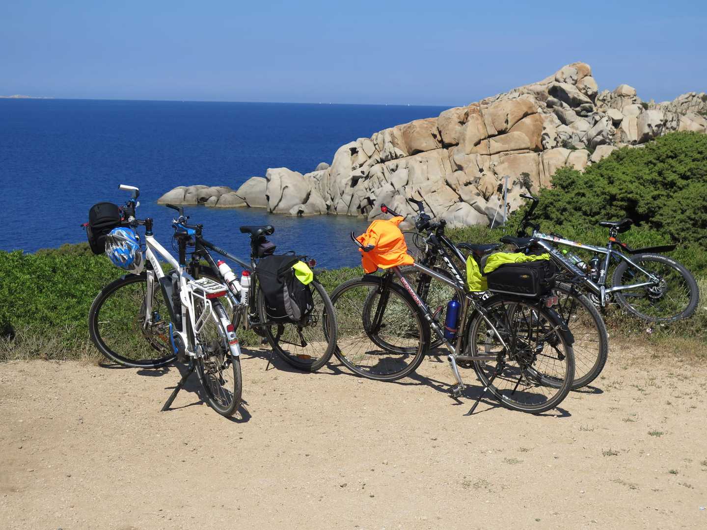 Radreise Italien Sardinien, mehrere Räder mit bunten Gepäckstücken an der sardischen Küste abgestellt mit Blick auf eine felsige Landzunge, die ins blaue Meer ragt