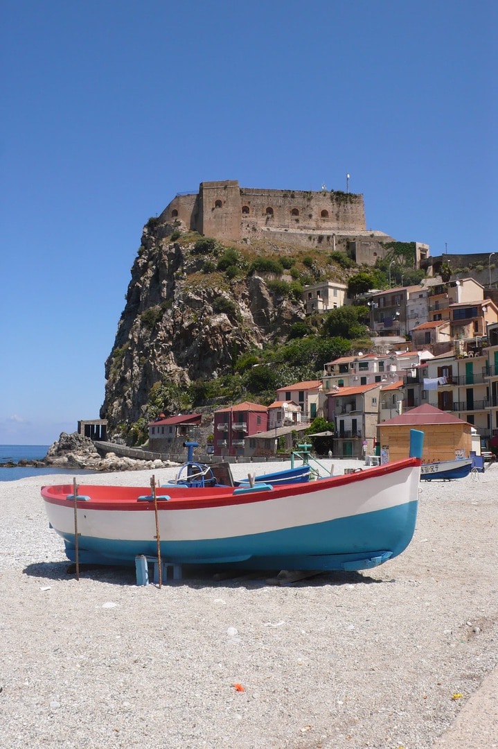 Radreise Italien Rom Sizilien, rot-weiss-blau gestrichenes Ruderboot am Strand einer kleinen sizilianischen Ortschaft mit einer Burgruine auf einem Felsvorsprung im Hintergrund