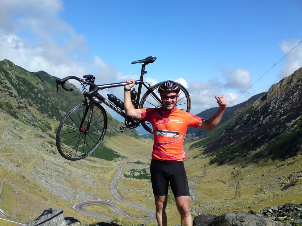 Rennradreise Rumänien Karpaten, Launer-Chef Paul Launer stolz ein Rennrad und die Arme hebend mit der soeben erklommenen Tranfagaraschen Hochstrasse im Hintergrund