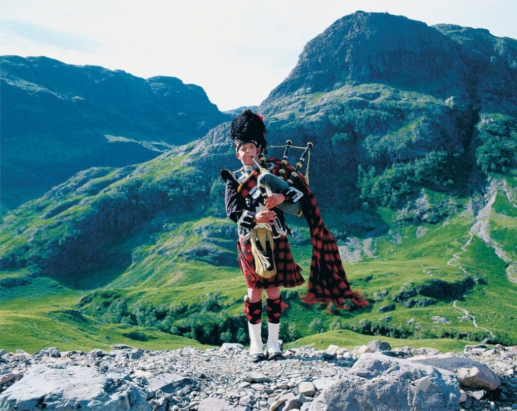 Radreise Großbritannien Schottland, junger Dudelsackspieler mit den rauhen und grünen Bergen der Highlands im Hintergrund